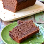 Tranches de cake au chocolat moelleux posées sur une assiette verte, avec le reste du gâteau au chocolat en arrière-plan.