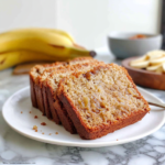 Tranches de recette banana bread moelleux servies sur une assiette blanche, avec des bananes mûres en arrière-plan.