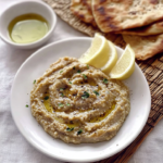 Creamy homemade caviar d'aubergine served with olive oil, lemon wedges, fresh herbs, and warm pita bread on a white plate.